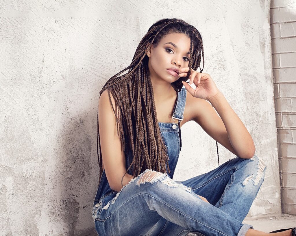 A woman with long braids sits against a textured, off-white wall, wearing blue denim overalls and ripped jeans, exuding a relaxed, thoughtful vibe.