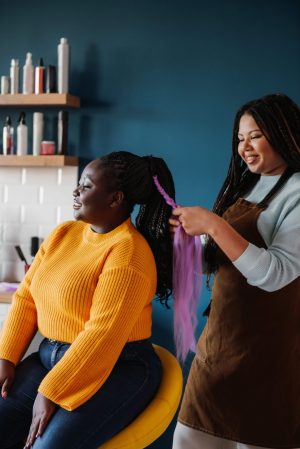 Two women are in a hair salon. One is seated, smiling, in a yellow sweater, while the other braids her hair with purple extensions. The mood is cheerful. Two women are in a hair salon. One is seated, smiling, in a yellow sweater, while the other braids her hair with purple extensions. The mood is cheerful.